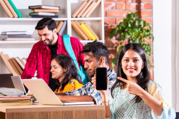 Group of four indian asian young college friends with backpack, books, laptop standing isolated on white, studying in library Indian asian college students in group using laptop computer pointing at blank or presenting or celebrating success, presenting blank placard, thumbs up success gesture, sitting on floor